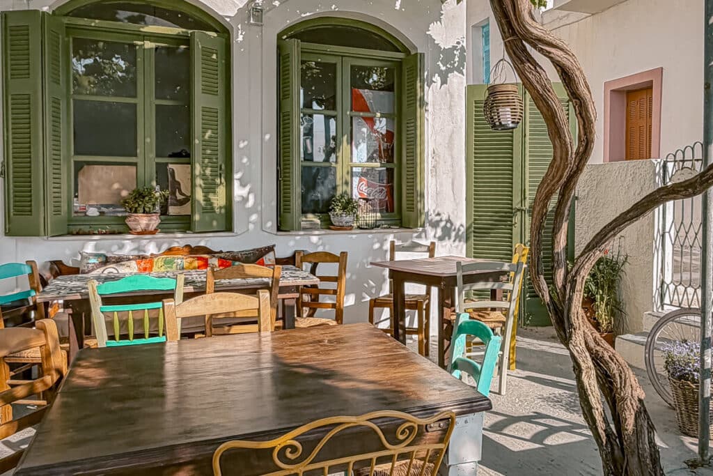 Shaded outdoor taverna in Andros with wooden tables, green shutters, colorful chairs, and a tree beside the seating area.