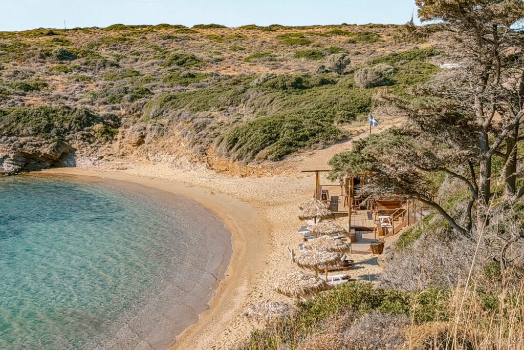 Sandy beach in Andros with clear turquoise water, straw umbrellas, a small beach bar, and rocky green hills behind it.