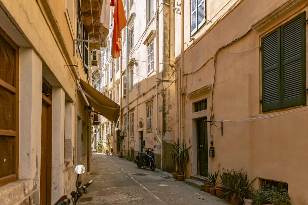 Narrow alley in Corfu Old Town with warm pastel buildings, shuttered windows, potted plants, and a scooter parked along the stone street.