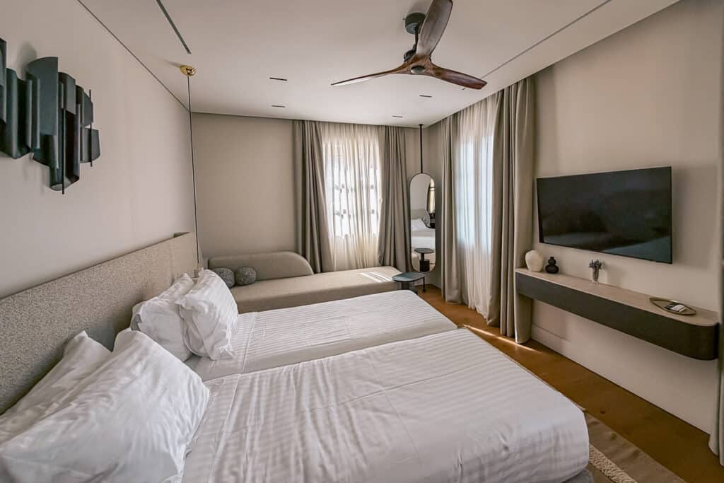 Modern hotel room at The Calliston in Corfu Old Town with a large bed, sofa, curtains, ceiling fan, and wall mounted TV.