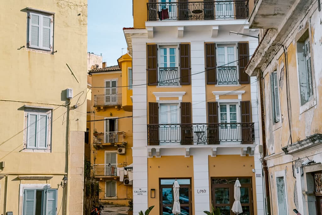 Exterior of The Calliston hotel in Corfu Old Town, with yellow and white balconies surrounded by pastel buildings and shuttered windows.