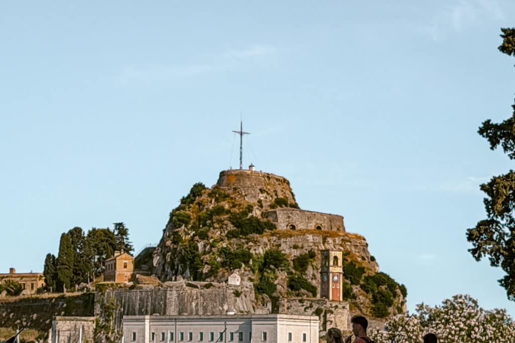 Stone walls of the Old Fortress in Corfu Town rising above trees, with a cross at the top and people walking below.