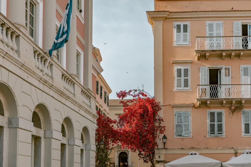 Greek flag, arched building, red flowers, and pastel balconies near Spianada Square in Corfu Old Town.