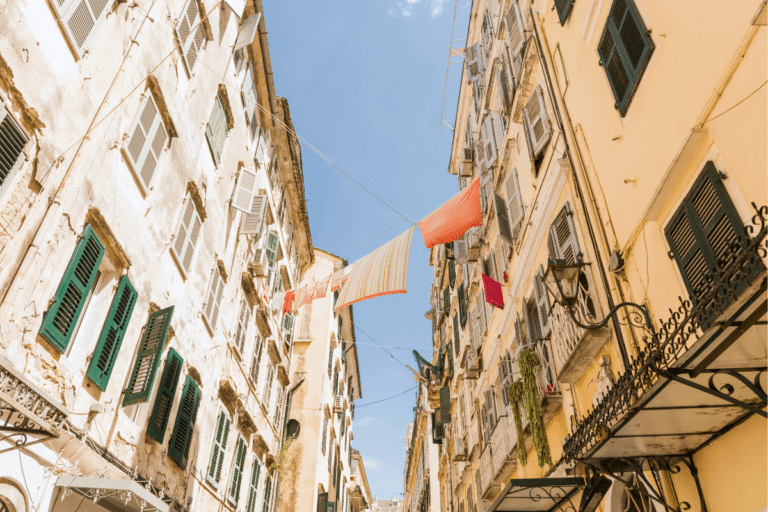 Narrow street in Corfu Old Town with tall pastel buildings, green shutters, balconies, and laundry hanging above the walkway on a sunny day to showcase things to do in Corfu Old Town.