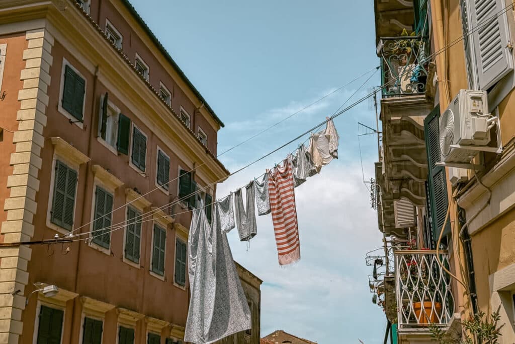 Laundry hanging between pastel buildings with green shutters and balconies on a narrow street one of the things to do in Corfu Old Town is to wander the streets.