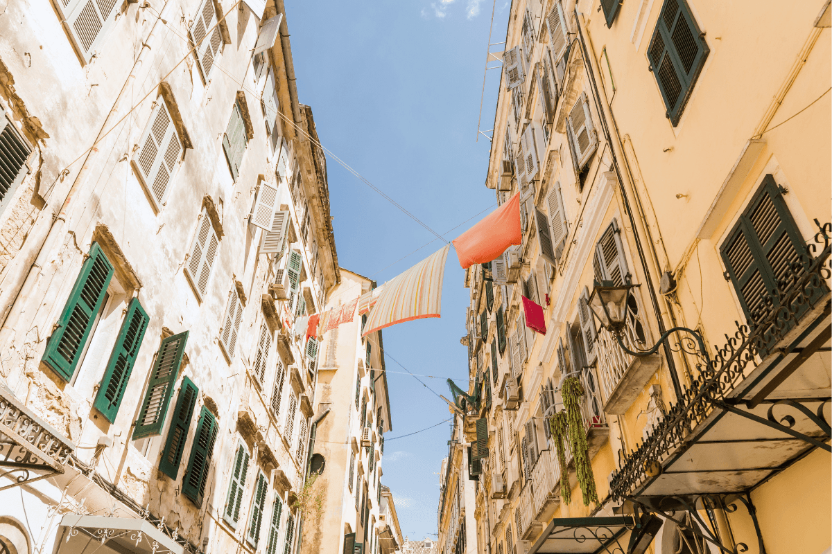 Narrow street in Corfu Old Town with tall pastel buildings, green shutters, balconies, and laundry hanging above the walkway on a sunny day to showcase things to do in Corfu Old Town.