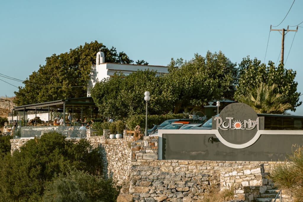 Exterior of Rotonda restaurant and cocktail bar in Naxos, set among trees and stone walls with outdoor seating and parked cars.