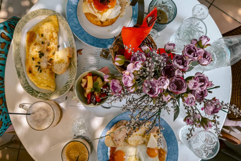 Overhead view of breakfast in Naxos with an omelet, eggs on toast, fruit, iced coffee, glassware, and a purple floral centerpiece.