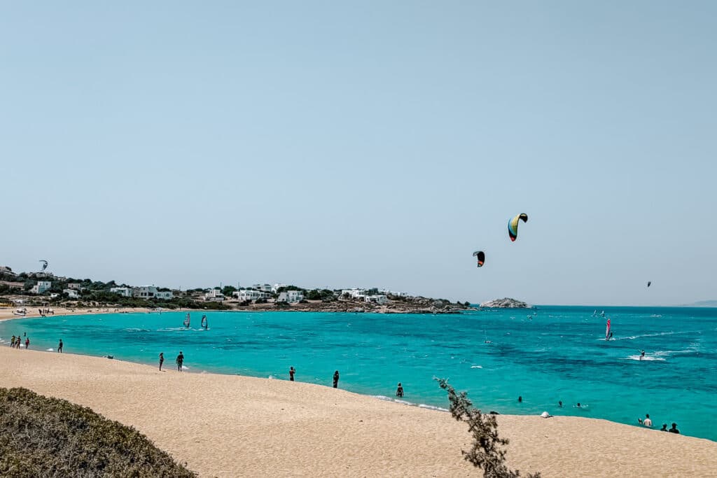 Mikri Vigla Beach in Naxos with turquoise water, a wide sandy shore, swimmers, windsurfers, and colorful kites in the sky.