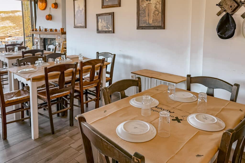 Interior of a traditional Naxos taverna with wooden tables, woven chairs, framed black and white photos, and mountain views through the windows.