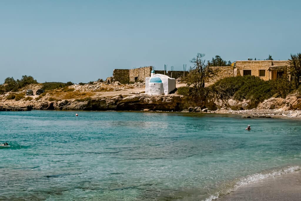 Small rocky cove in Naxos with clear turquoise water, swimmers, a white chapel with a blue roof, and rustic stone buildings along the shore.