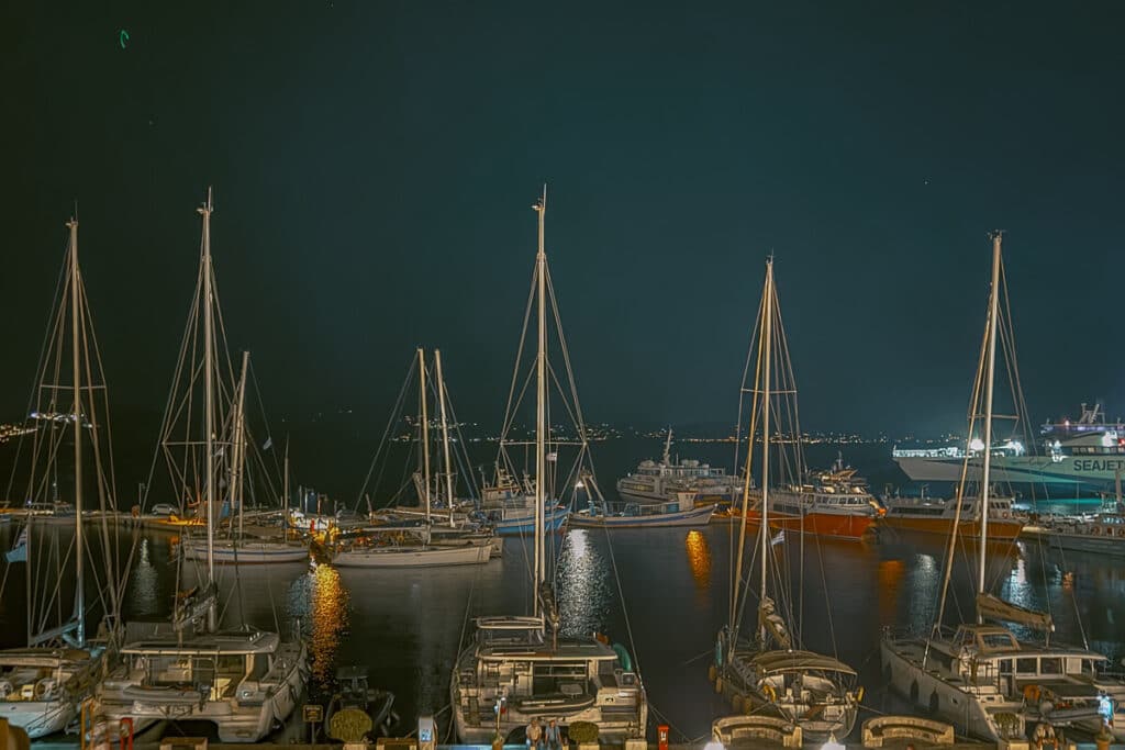 Sailboats and ferries docked in Naxos harbor at night, with lights reflecting across the dark water.