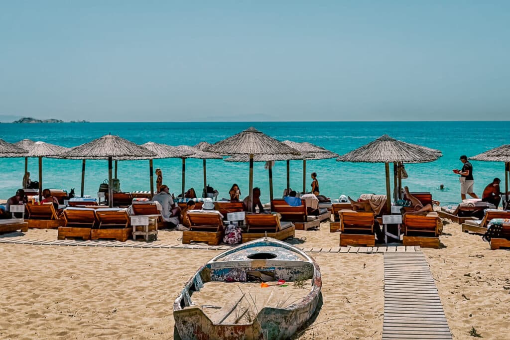 Organized beach in Naxos with wooden sunbeds, straw umbrellas, swimmers in turquoise water, and a weathered fishing boat resting on the sand.