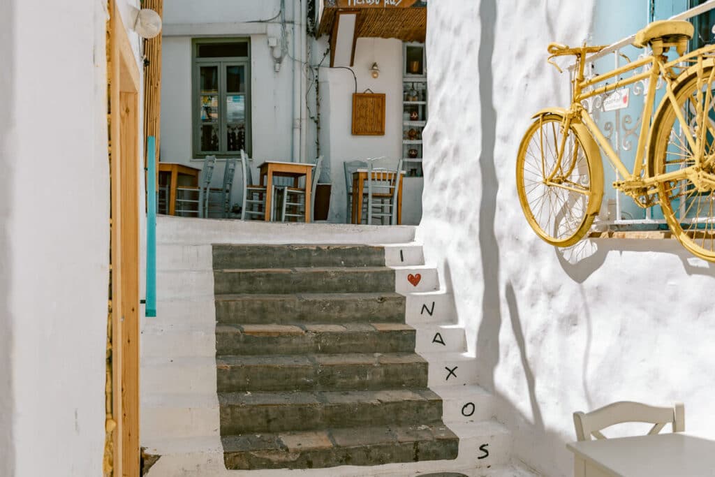 Stone steps in Naxos Old Town leading to a small café, with whitewashed walls, gray chairs, a yellow bicycle, and “I love Naxos” painted beside the stairs.