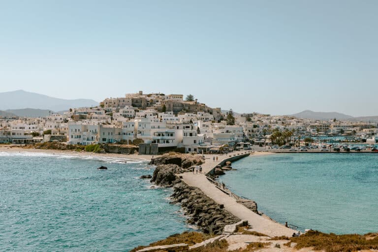 Things to do in Naxos, Greece, with a panoramic view of Chora’s whitewashed buildings, hilltop Kastro, waterfront harbor, and stone walkway surrounded by turquoise sea.