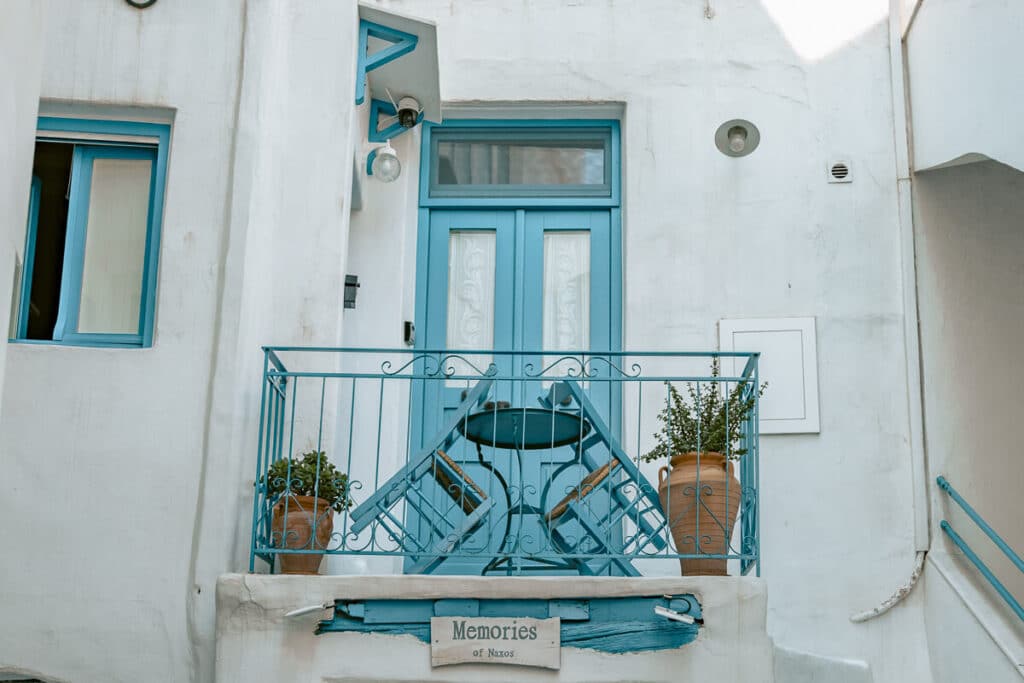 Small balcony in Naxos Old Town with turquoise doors, matching railings, café chairs, and terracotta plant pots against whitewashed walls.