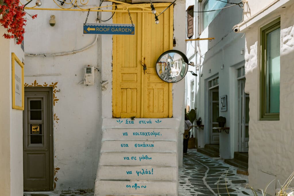 Narrow lane in Naxos Old Town with a yellow door, whitewashed steps painted with Greek words, and a sign pointing toward a roof garden.