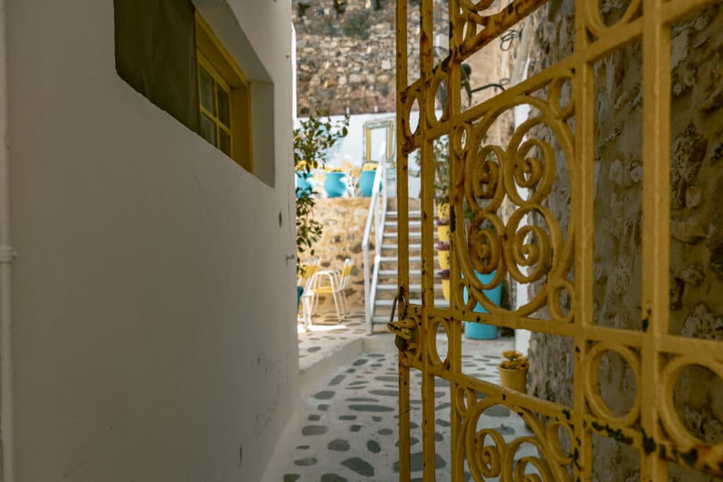 Yellow wrought iron gate opening into a narrow stone paved courtyard in Naxos Old Town, with whitewashed walls, steps, and turquoise seating beyond.