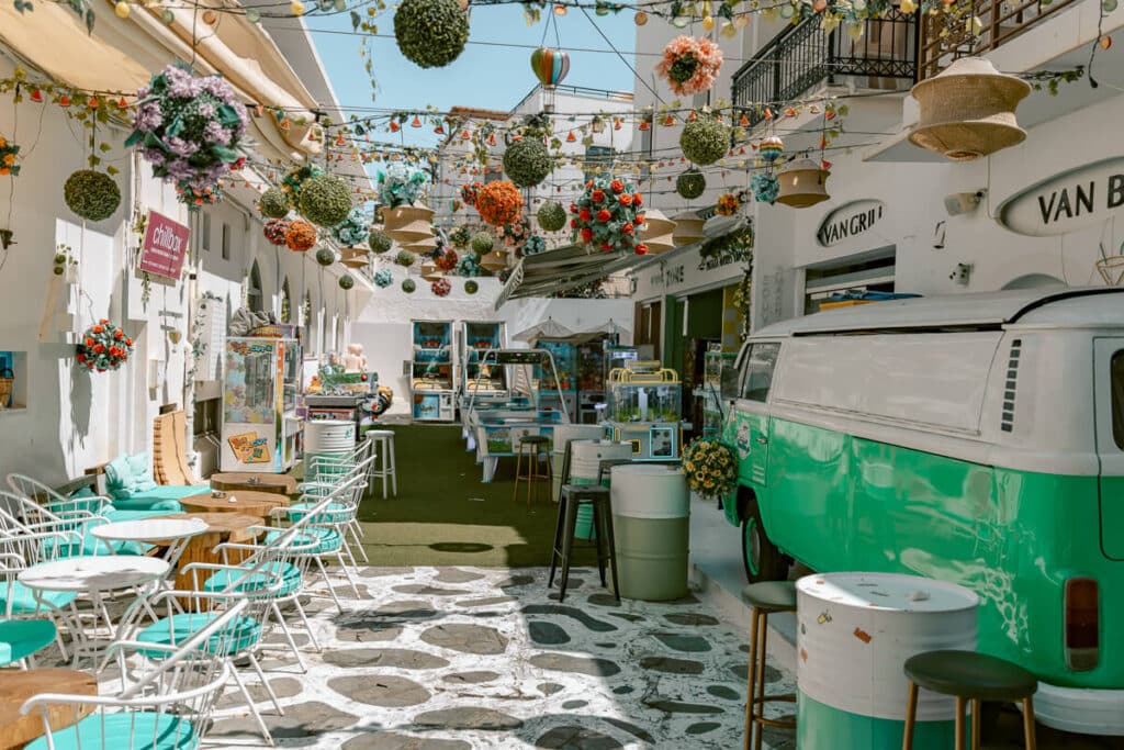 Colorful café courtyard in Naxos Old Town decorated with hanging flowers, string lights, turquoise seating, arcade games, and a green vintage van.