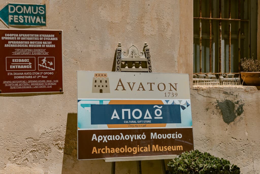 Signs for the Archaeological Museum of Naxos, Avaton, and a cultural gift shop displayed on a sunlit stone wall in the Kastro.