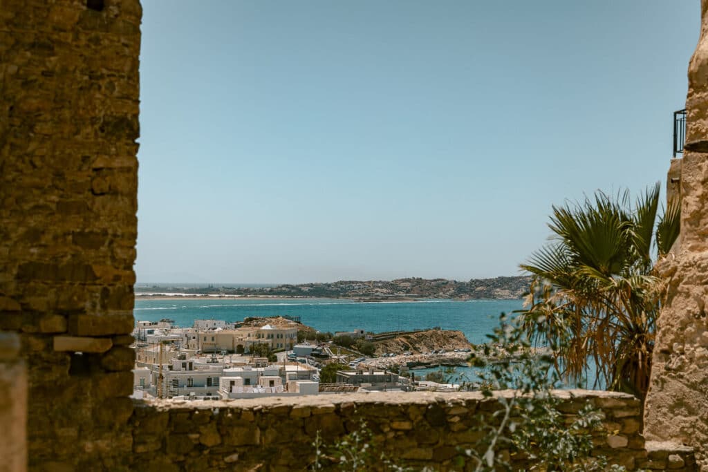 View from the Kastro in Chora over whitewashed buildings, the blue Aegean Sea, rocky coastline, and palm trees framed by old stone walls.
