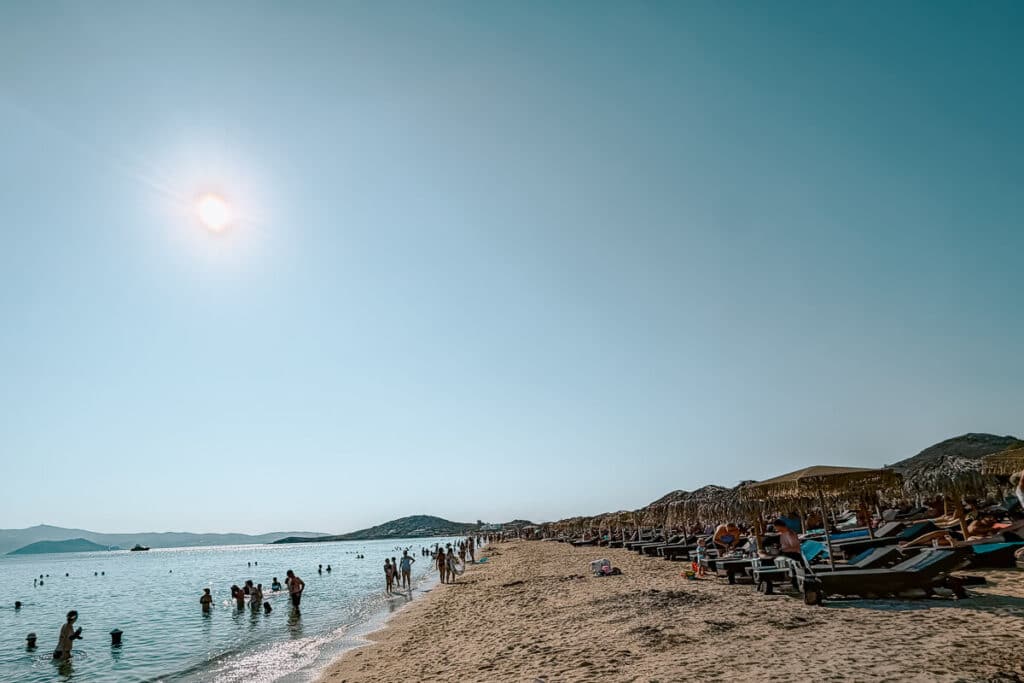 Busy sandy beach in Naxos with rows of sunbeds and straw umbrellas, people swimming in calm shallow water, and the afternoon sun overhead.