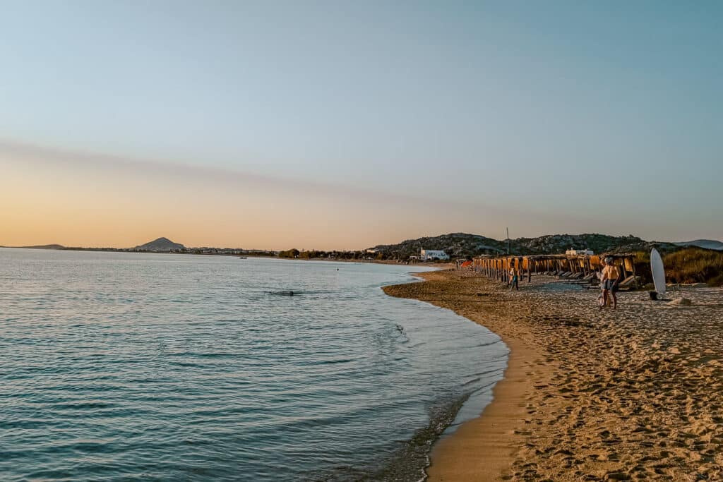 Quiet sandy beach in Naxos at sunset with calm water, rows of straw umbrellas, a paddleboard, and low hills in the distance.