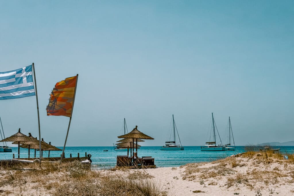 Sandy beach in Naxos with straw umbrellas, Greek and orange flags blowing in the wind, and sailboats anchored in the blue sea.