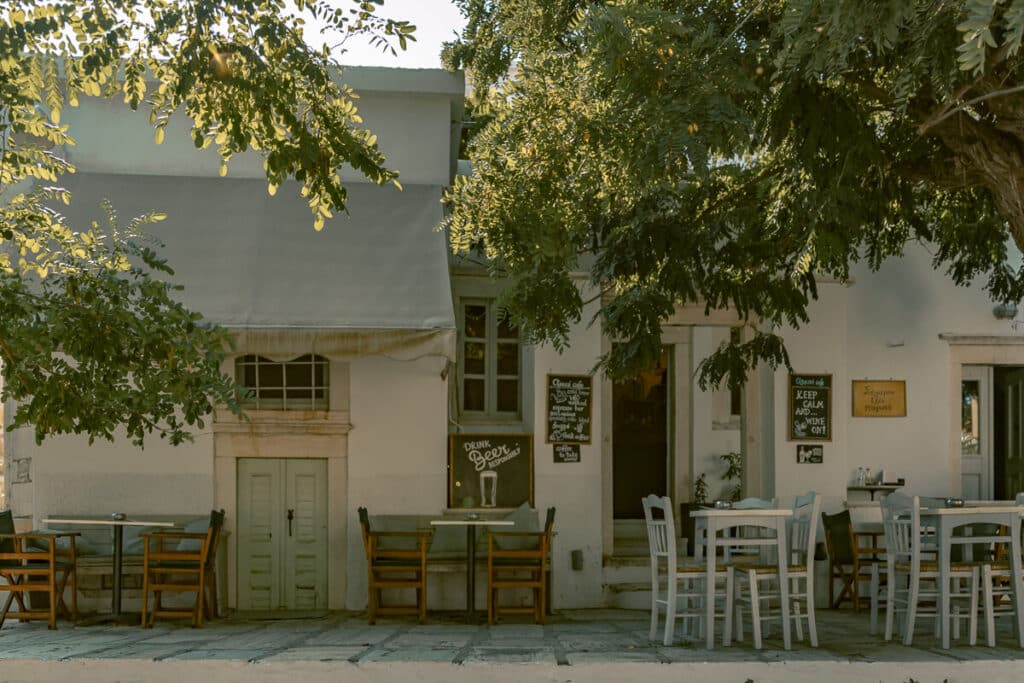 Shaded outdoor taverna in a Naxos mountain village with white tables, wooden chairs, chalkboard signs, and leafy trees overhead.