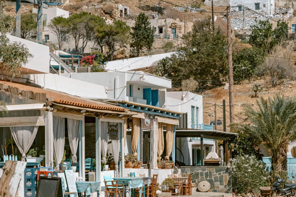 Outdoor taverna in Serifos with turquoise tables and chairs, whitewashed buildings, flowering plants, and a dry hillside in the background.