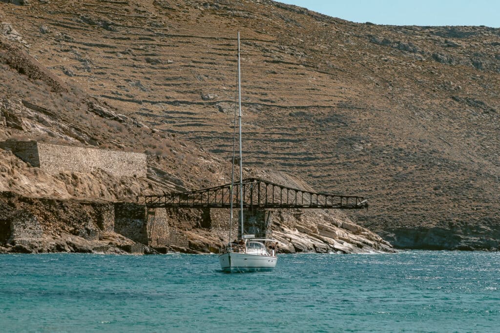 Sailboat floating in turquoise water off the coast of Serifos, with old mining structures and rugged terraced hills along the shoreline.