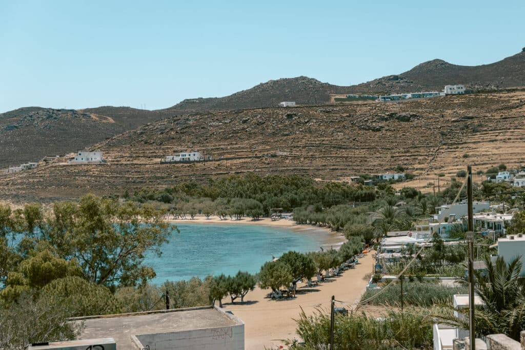 Livadakia Beach in Serifos with turquoise water, a sandy shoreline lined with shady trees, white buildings, and rugged hills in the background.
