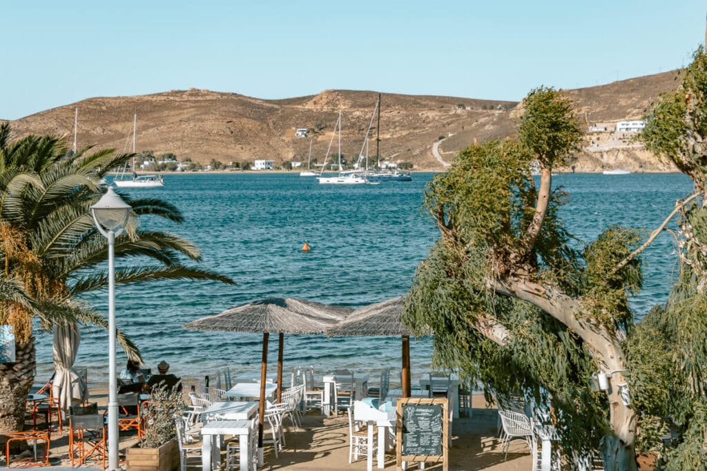 Waterfront café with tables and umbrellas overlooking blue water and sailboats in Livadi, Serifos, with dry hills in the background.