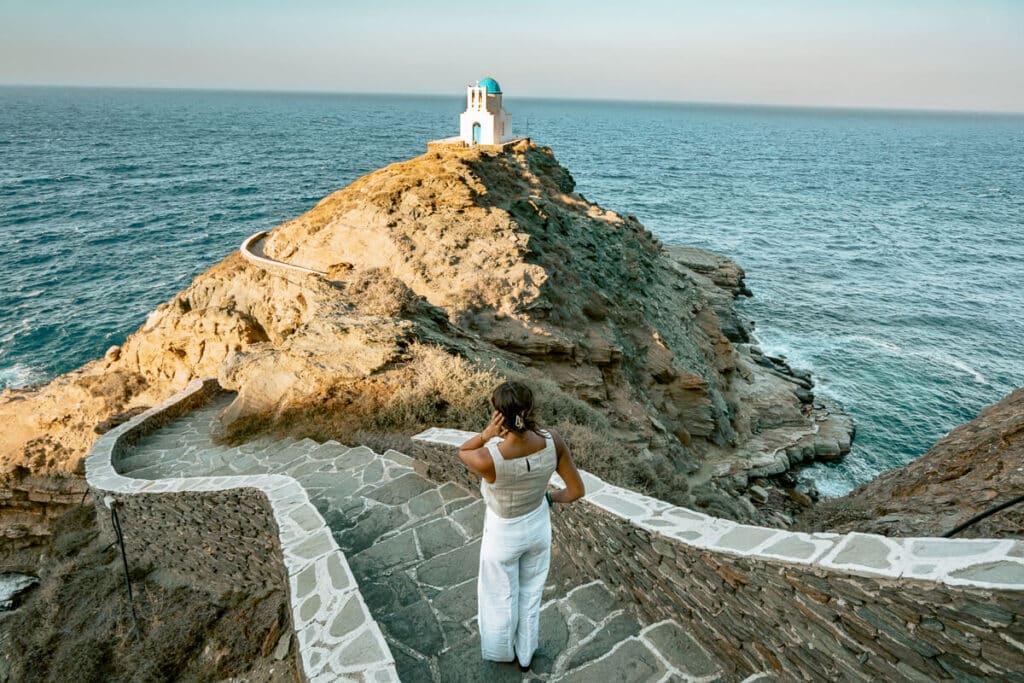 Me in white standing on a stone path overlooking the Church of the Seven Martyrs on a rocky peninsula surrounded by the Aegean Sea in Sifnos.