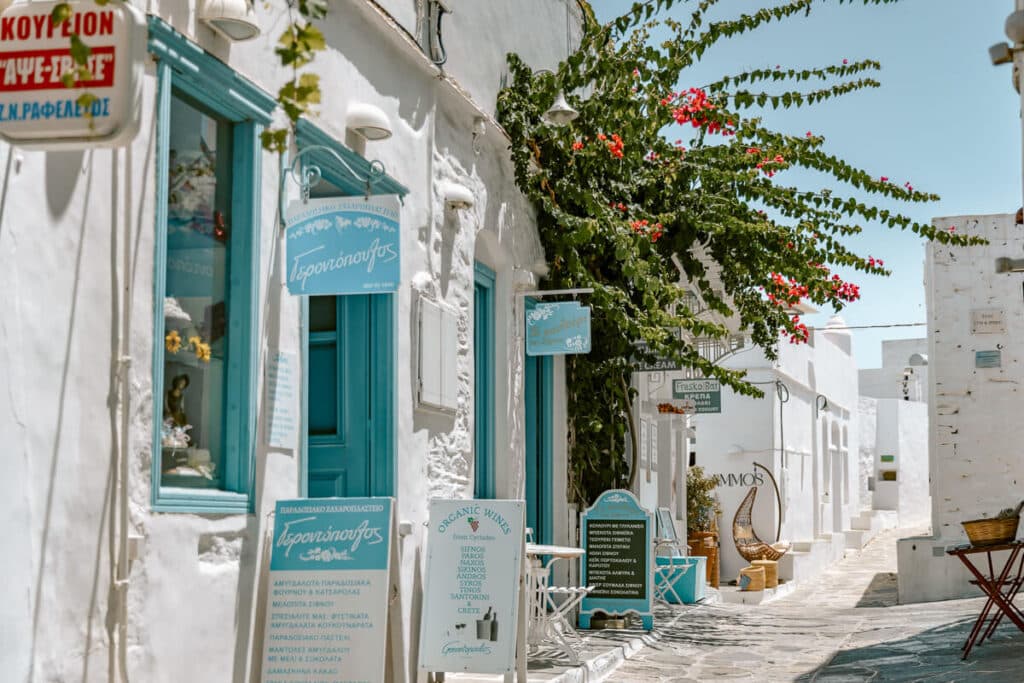 Whitewashed pedestrian street in Sifnos lined with turquoise doors, shop signs, outdoor seating, and red bougainvillea in a village in Sifnos.