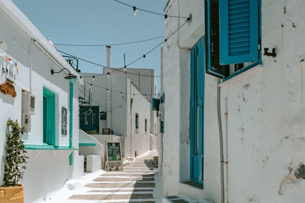 Narrow pedestrian street lined with whitewashed buildings, turquoise doors and blue shutters beneath string lights in Chora, Serifos.