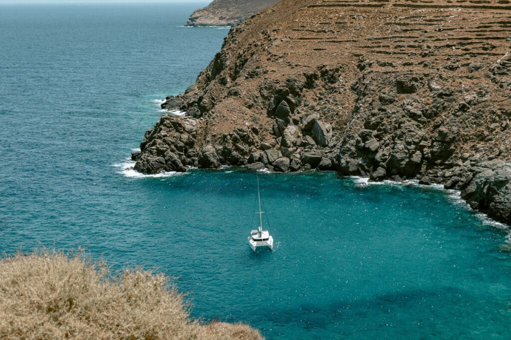White sailboat anchored in turquoise Aegean water beneath the rugged, rocky coastline of Sifnos.