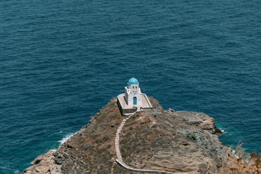 White Church of the Seven Martyrs with a blue dome perched on a rocky peninsula surrounded by the deep blue Aegean Sea in Sifnos, Greece.