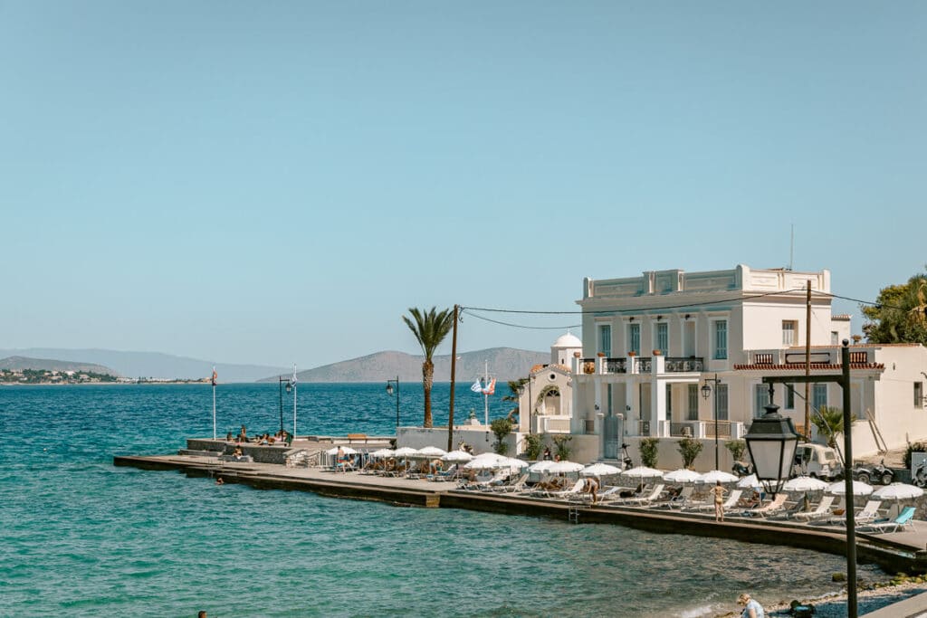 White waterfront hotel with sun loungers and umbrellas beside the turquoise sea in Spetses, Greece.
