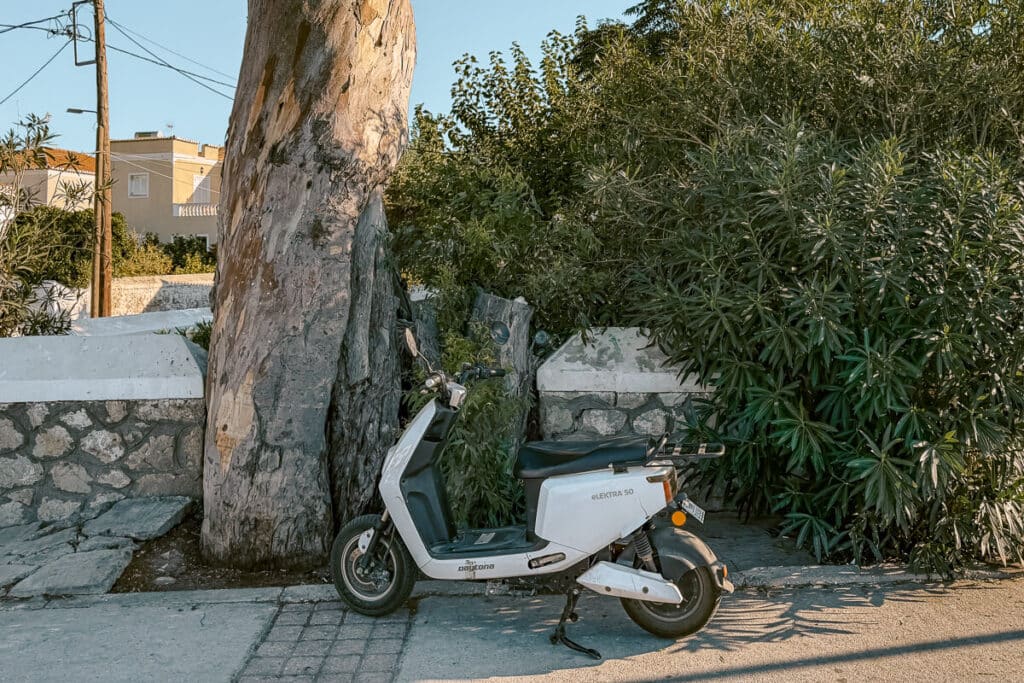 White scooter parked beside a large tree and stone wall on a quiet street in Spetses, Greece.
