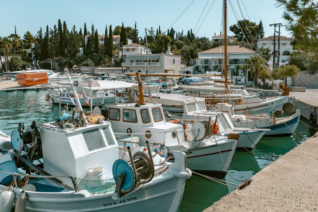 Small fishing boats and sailboats docked in the turquoise water of a harbor in Spetses, with white homes and trees along the waterfront.