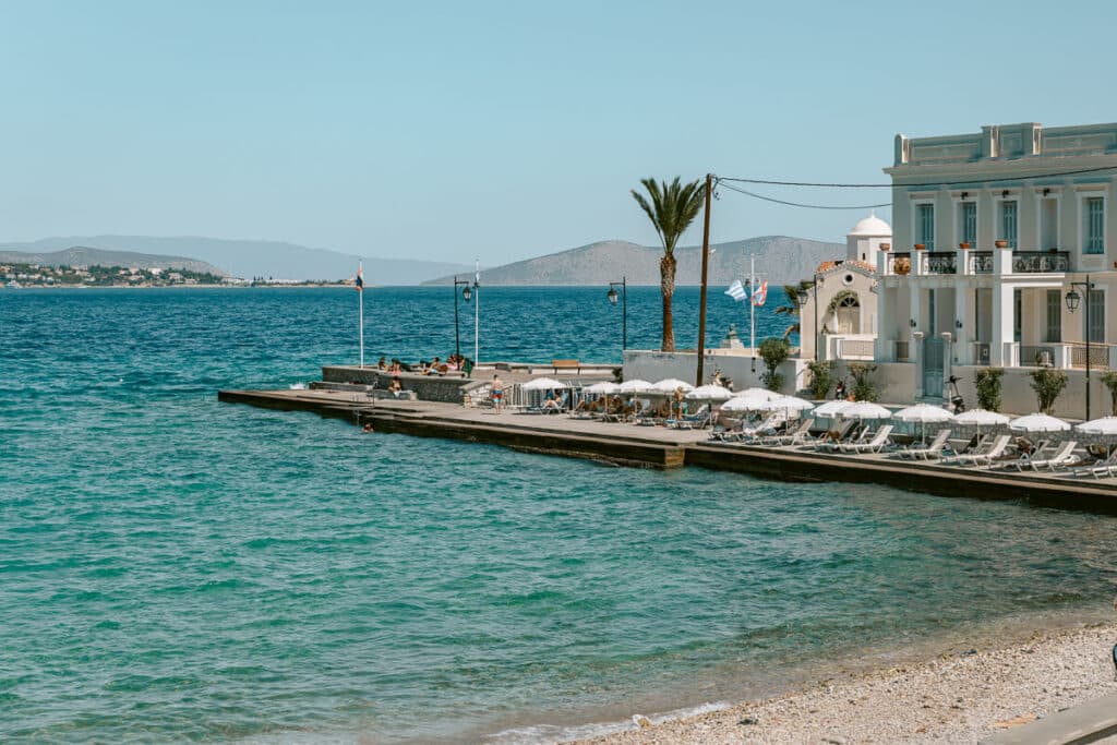 Agios Mamas Beach in Spetses with clear turquoise water beside a white waterfront building, sun loungers, umbrellas, and people swimming.
