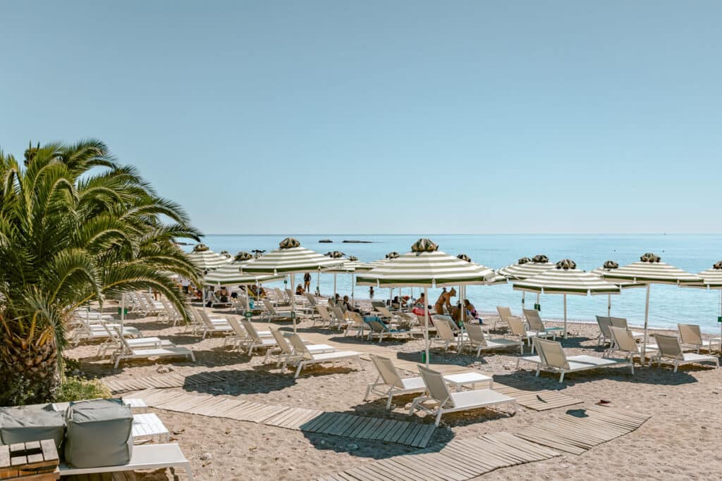 Agia Marina Beach in Spetses with white sun loungers, green and white striped umbrellas, palm trees, and people swimming in the calm blue sea.