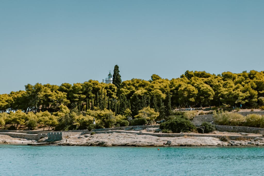 Spetses Lighthouse partially visible above a pine-covered coastline, with rocky shore and turquoise sea in the foreground.