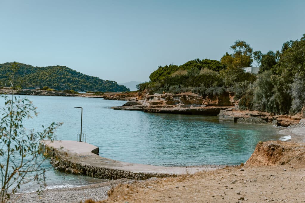 Quiet rocky cove in Spetses with clear turquoise water, a small pebble beach, concrete pier, and tree-covered coastline.
