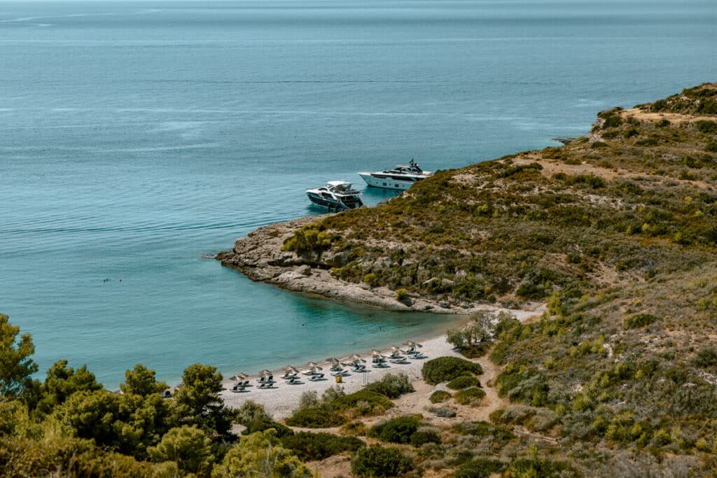 Secluded cove in Spetses with a pebble beach, rows of sun loungers and umbrellas, turquoise water, and yachts anchored beside the rocky coastline.