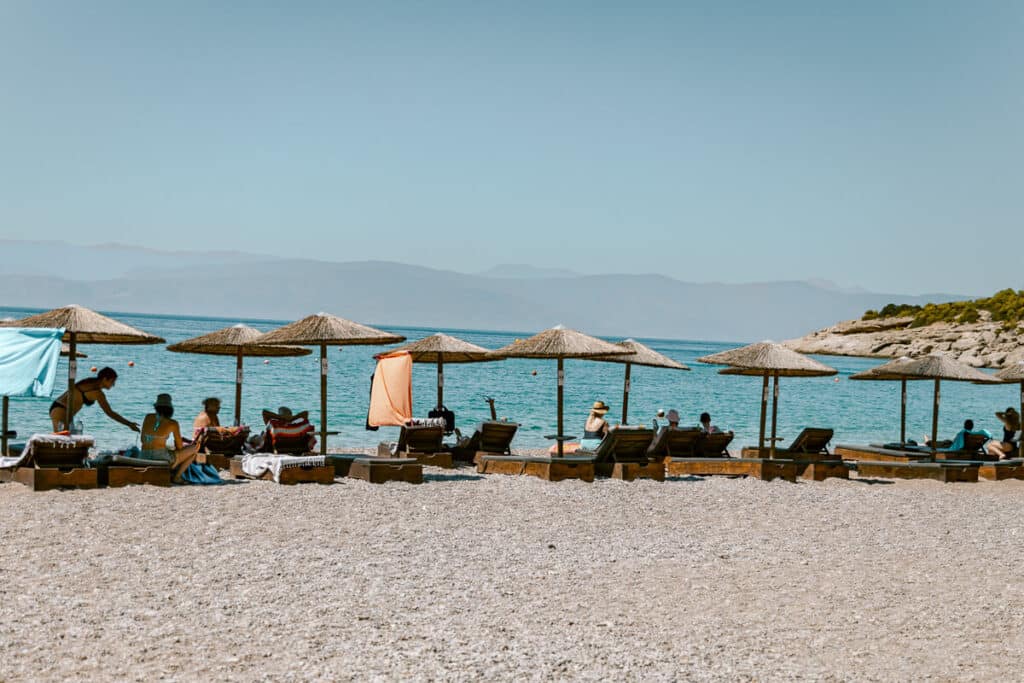 Pebble beach in Spetses with straw umbrellas and sun loungers overlooking turquoise water and distant mountains.