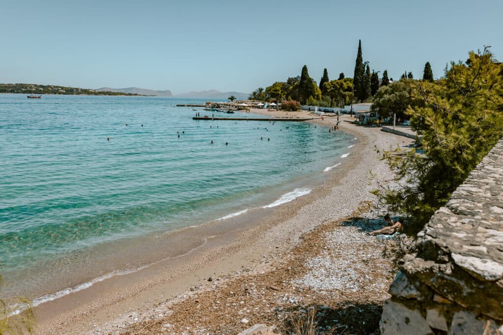 Curving pebble beach in Spetses with clear turquoise water, swimmers in the sea, and pine trees lining the shore.