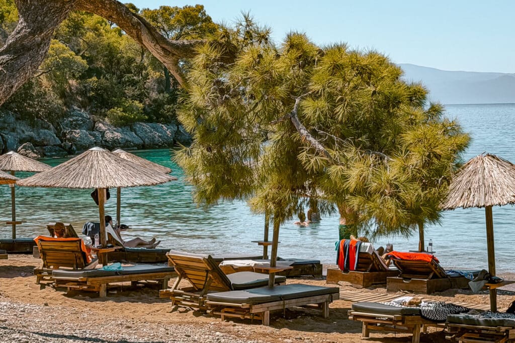 Spetses beach with wooden sun loungers and straw umbrellas beneath pine trees, overlooking clear turquoise water in a sheltered cove.