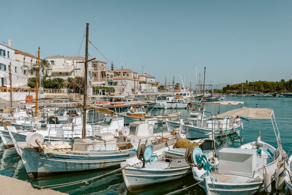 Traditional fishing boats docked in the turquoise water of Spetses harbor, with white waterfront buildings and sailboats in the background.